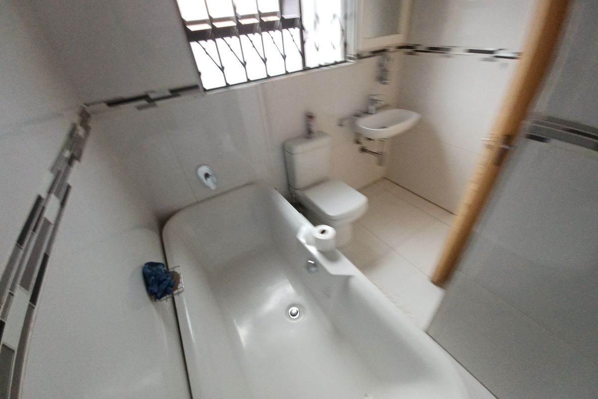 A functional bathroom featuring a bathtub, wall-mounted basin, and toilet with neutral tiled finishes and security bars on the window.