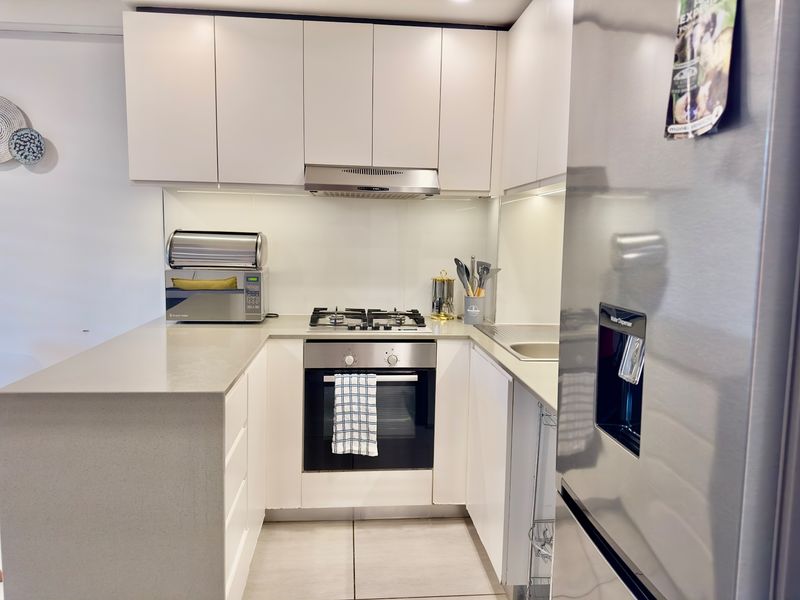 Modern compact kitchen featuring white cabinetry, stone benchtops, gas cooktop, built-in oven, and a sleek glass splashback.