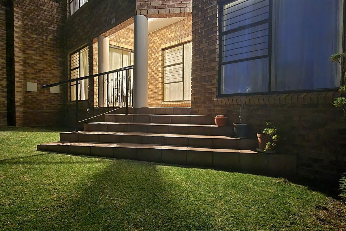 Night view of a face brick apartment exterior featuring a tiled patio entrance with wrought iron railings and security-fitted windows overlooking a lawn.