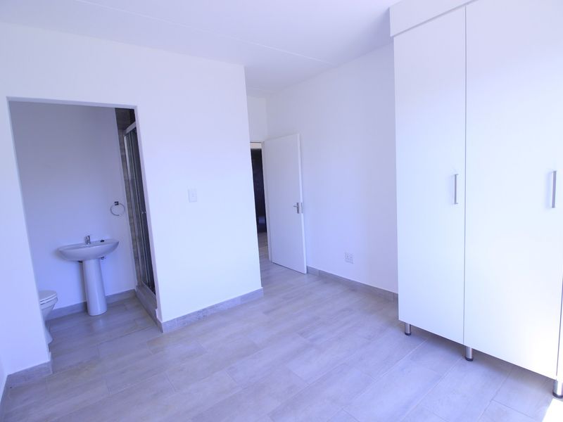 A bright, empty bedroom featuring tiled flooring, a large built-in wardrobe, and an attached en-suite bathroom with a pedestal basin and shower.