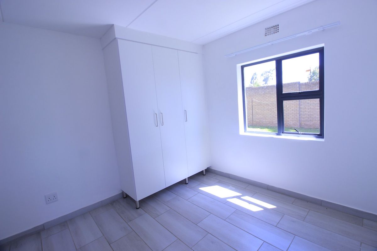 A bright, empty bedroom featuring neutral tiled flooring, a built-in wardrobe, and a window with views of the exterior brick wall.
