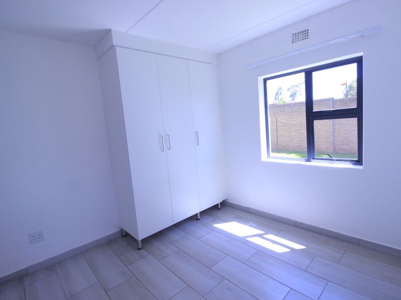A bright, empty bedroom featuring neutral tiled flooring, a built-in wardrobe, and a window with views of the exterior brick wall.