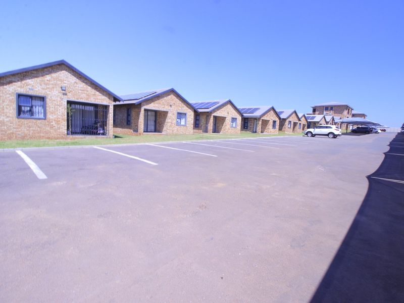 A row of face brick apartment units featuring solar panels, security gates, and a large paved parking area with carports.