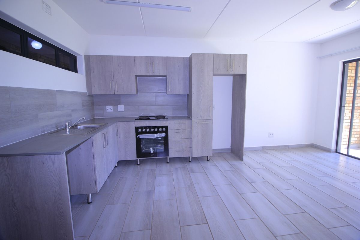 Modern open-plan kitchen featuring light wood-grain cabinetry, built-in oven with gas hob, stainless steel sink, and neutral tiled flooring throughout.