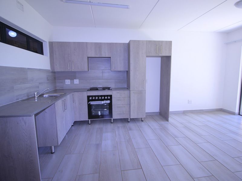 Modern open-plan kitchen featuring light wood-grain cabinetry, built-in oven with gas hob, stainless steel sink, and neutral tiled flooring throughout.