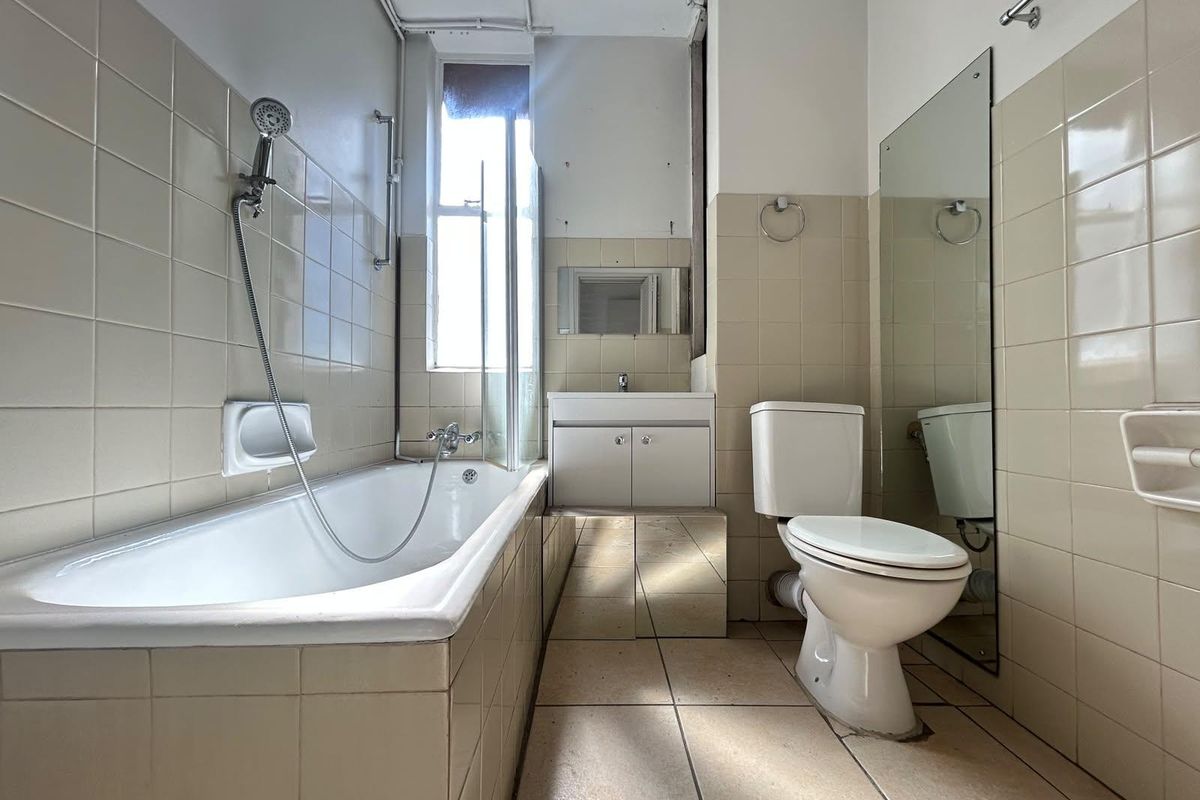 A dated bathroom featuring a bathtub with shower mixer, vanity unit with basin, and toilet, finished with neutral wall and floor tiling.