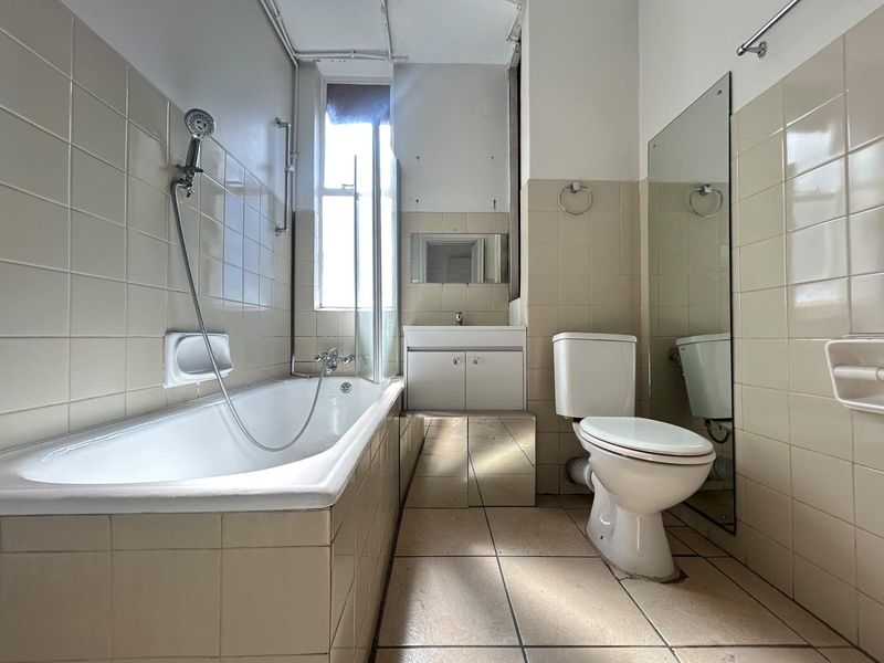 A dated bathroom featuring a bathtub with shower mixer, vanity unit with basin, and toilet, finished with neutral wall and floor tiling.