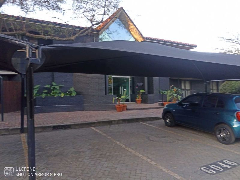 Exterior view of the apartment building entrance featuring a paved parking area with a shade sail and dark brick facade.