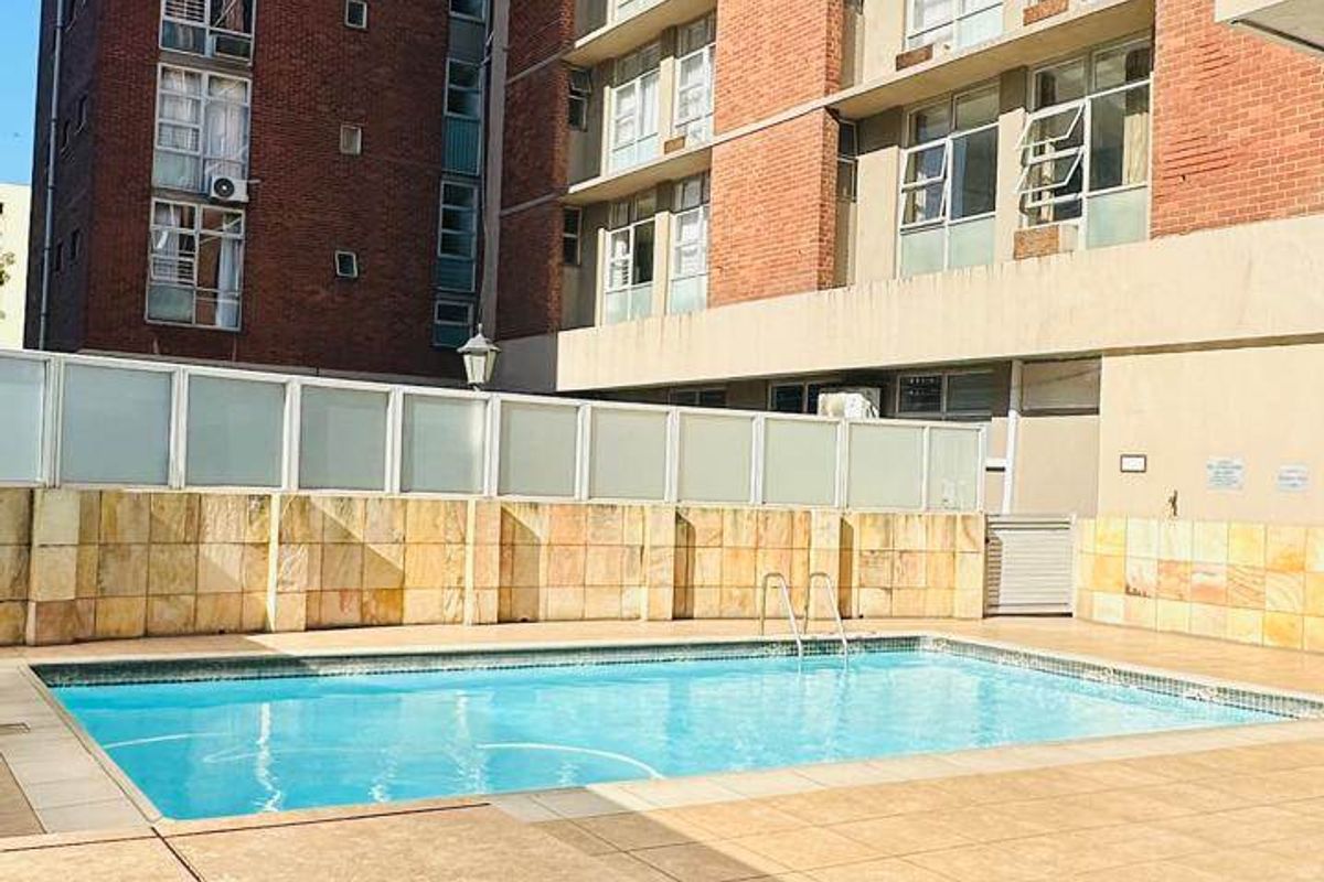 A well-maintained communal swimming pool area featuring stone-clad walls, tiled decking, and a pool ladder, set against a multi-story apartment building.