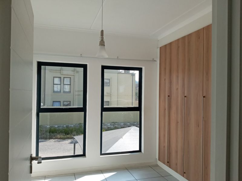 A bright, modern bedroom featuring neutral tiled flooring, sleek built-in wooden cupboards, and large aluminium-framed windows providing ample natural light.