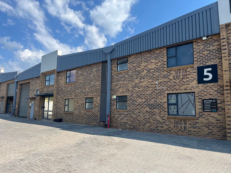Front elevation of a modern industrial unit featuring face brick construction, metal roof cladding, aluminium windows, and a paved driveway.