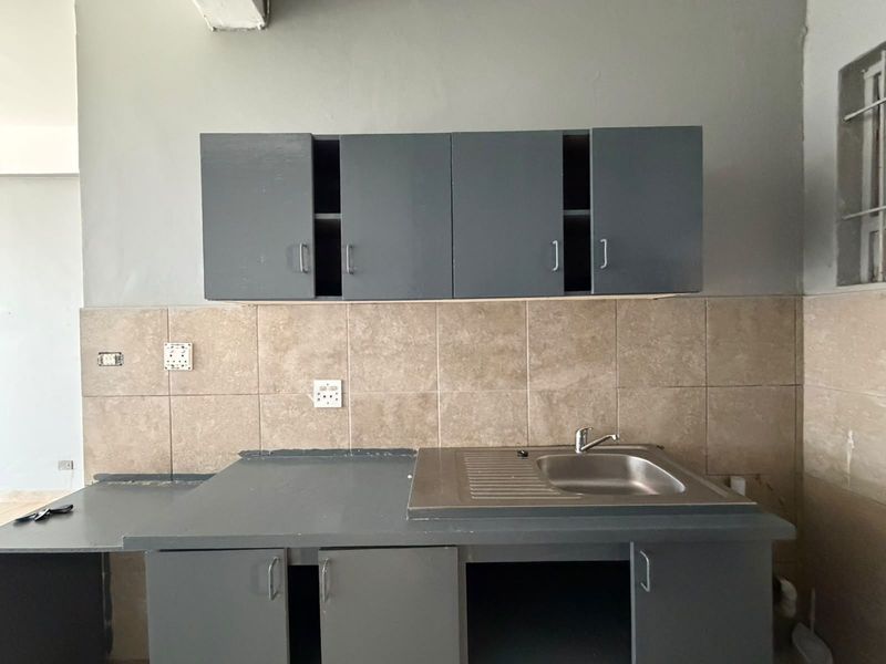 A basic kitchen area featuring built-in grey cabinetry, a stainless steel sink, and a tiled backsplash, currently in need of cosmetic updates and finishing.