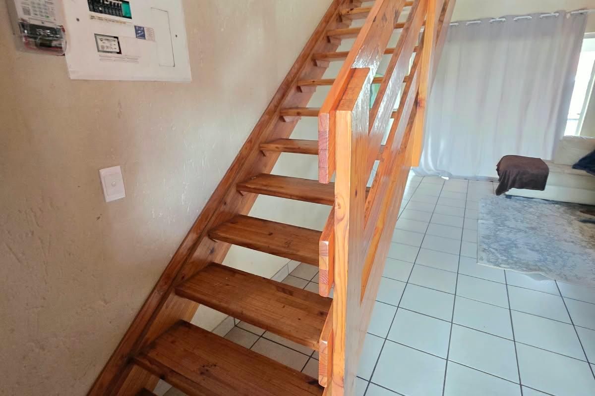 Open plan living area featuring a wooden staircase leading to a mezzanine level, with tiled flooring and a wall-mounted electrical distribution board.