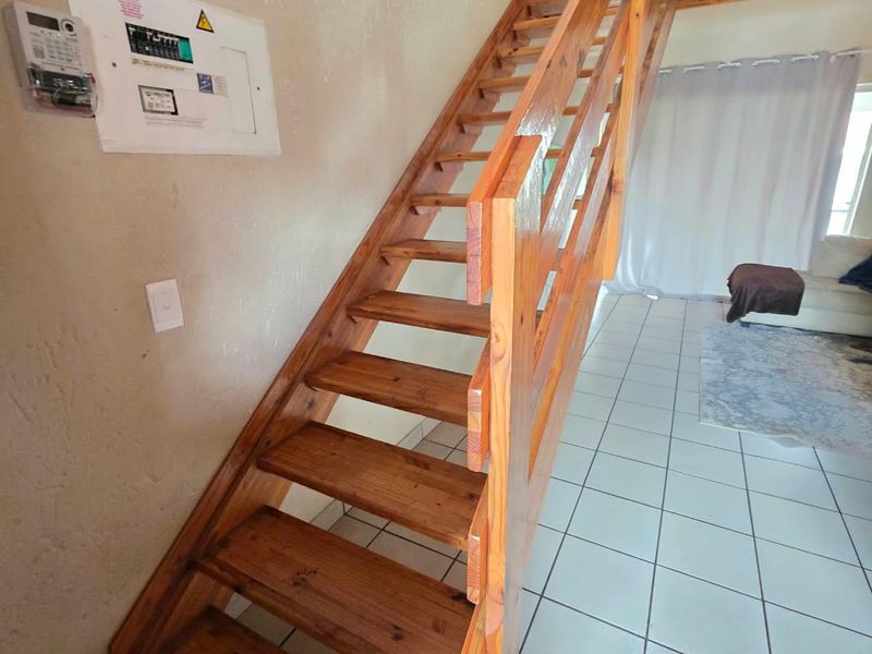 Open plan living area featuring a wooden staircase leading to a mezzanine level, with tiled flooring and a wall-mounted electrical distribution board.