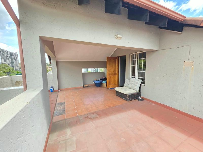A covered balcony area featuring tiled flooring, a timber entrance door, and exterior wall lighting, offering a functional outdoor space in need of minor maintenance.