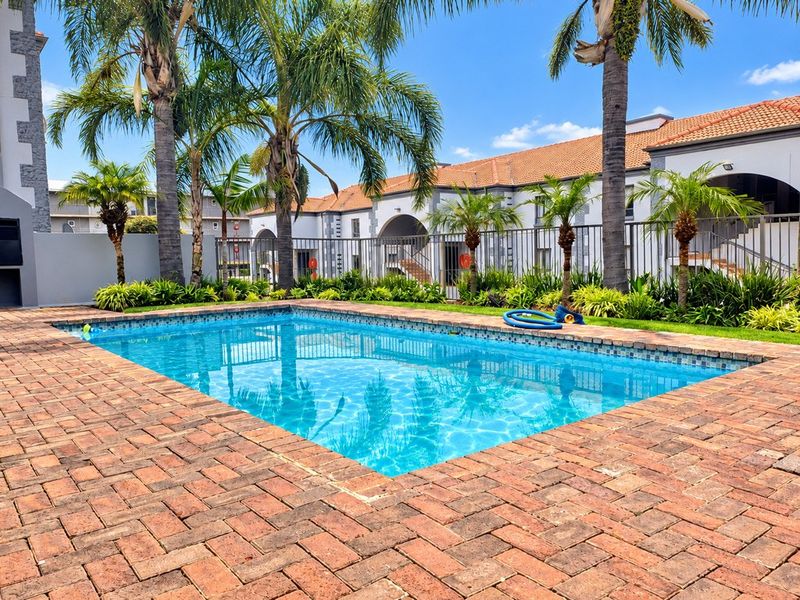 A well-maintained outdoor swimming pool area featuring brick paving, established palm trees, and secure metal perimeter fencing within a residential apartment complex.