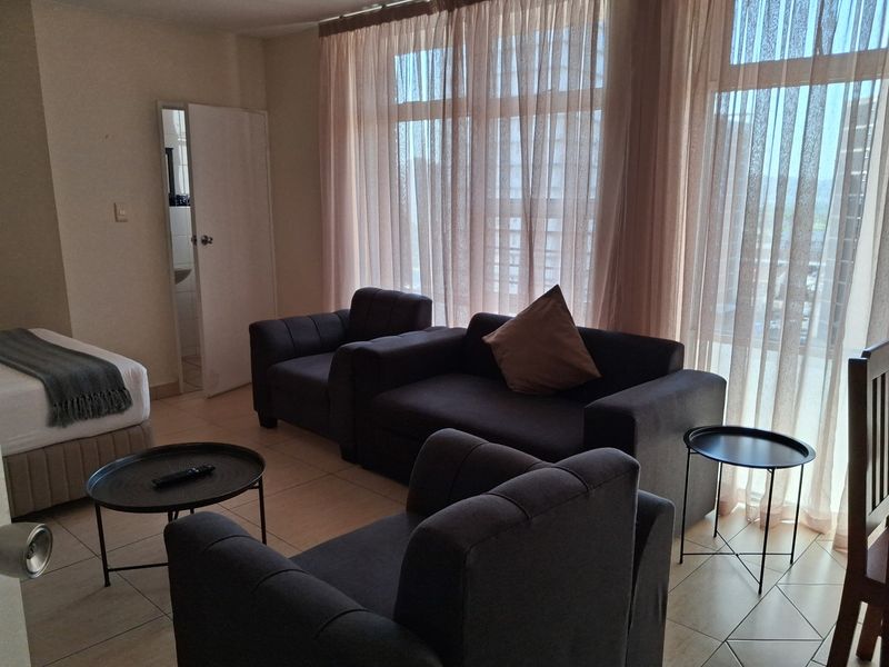 Open plan living area featuring tiled flooring and large windows with sheer curtains, providing ample natural light and access to an adjacent room.