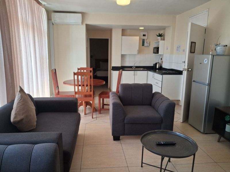 An open-plan living and kitchen area featuring tiled flooring, a compact kitchen with built-in cabinetry, and a wall-mounted air conditioning unit.