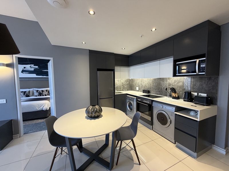 Modern open-plan kitchen featuring sleek dark and white cabinetry, stone-look backsplash, integrated appliances, and tiled flooring, seamlessly connecting to the adjacent bedroom.