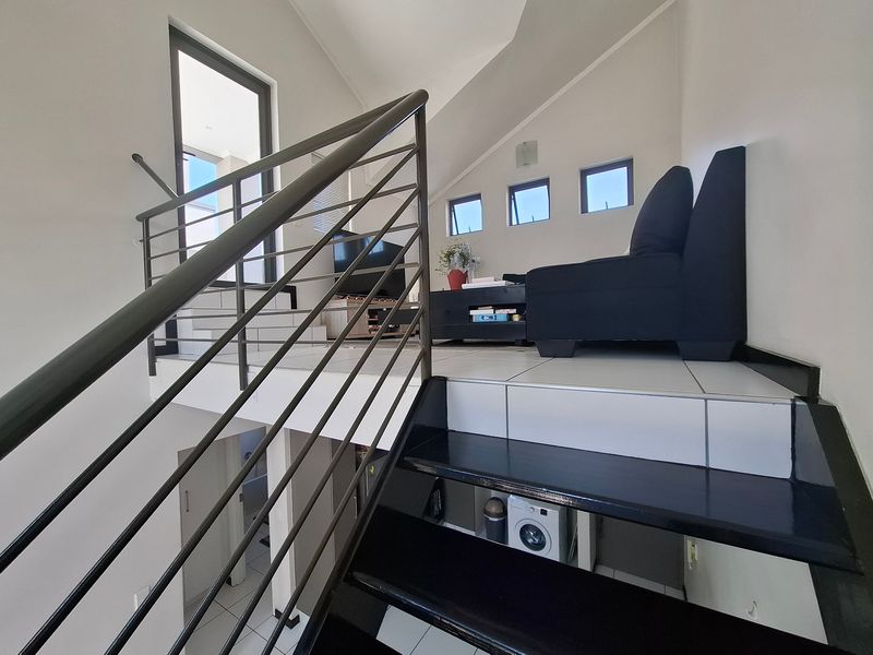 A bright living area featuring a vaulted ceiling, tiled flooring, and a modern staircase with a metal balustrade leading to an upper level.