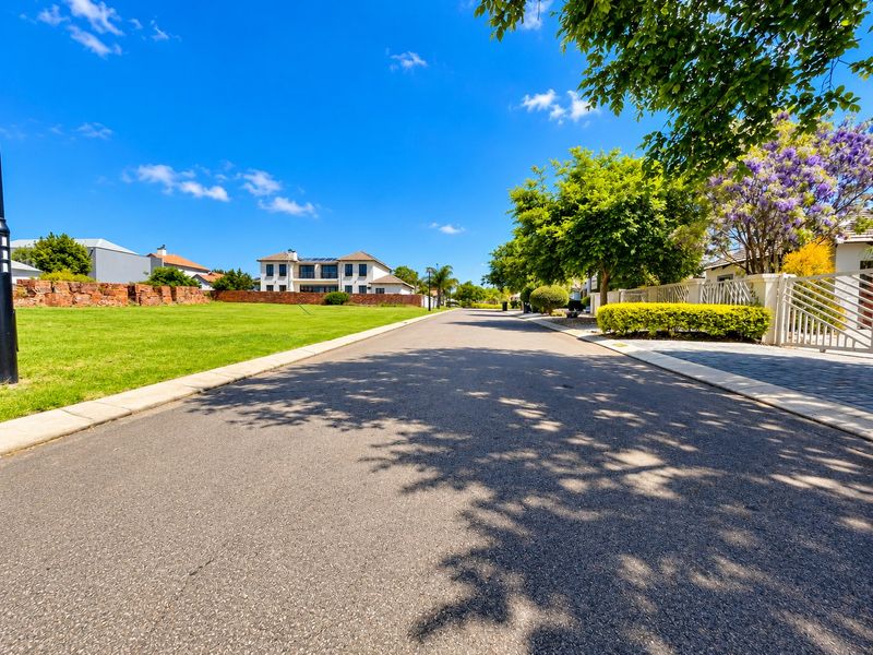 A clear street view of a well-maintained residential estate road featuring paved surfaces, manicured verges, and established stone boundary walls.