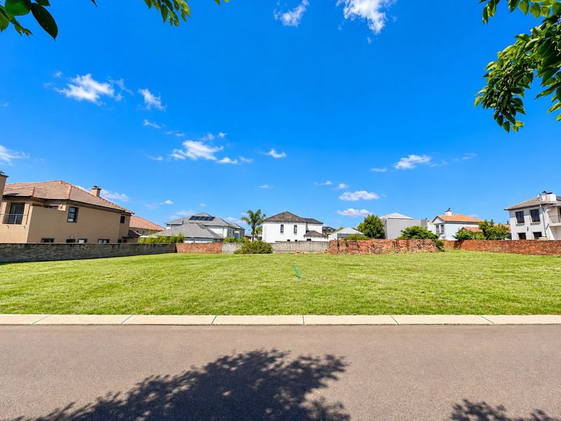 A vacant residential plot featuring a level grassed area, paved road frontage, and existing boundary walls in a developed neighborhood.