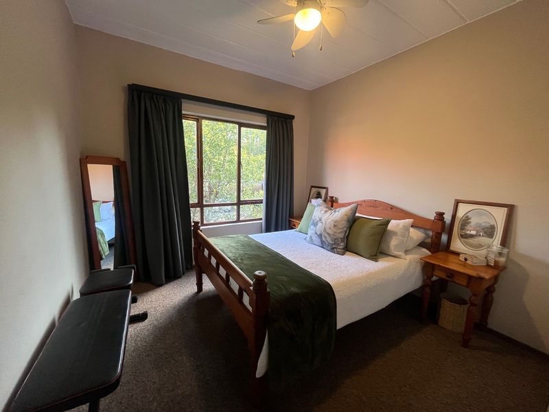 A cozy bedroom featuring carpeted flooring, a ceiling fan, and a large window that provides natural light and a view of the greenery outside.