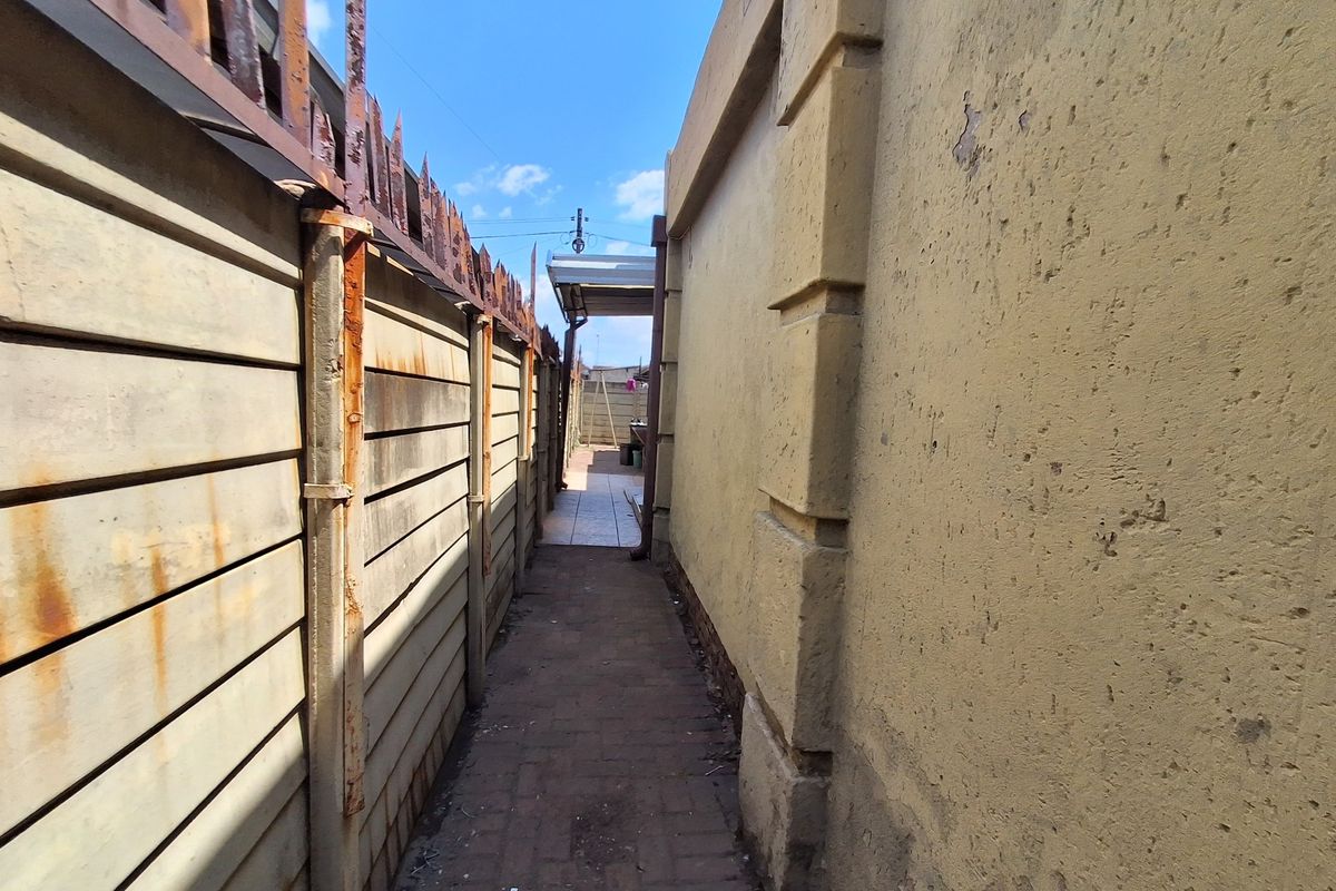 A narrow side passage featuring a paved walkway between a precast concrete boundary wall with security spikes and the main building exterior.