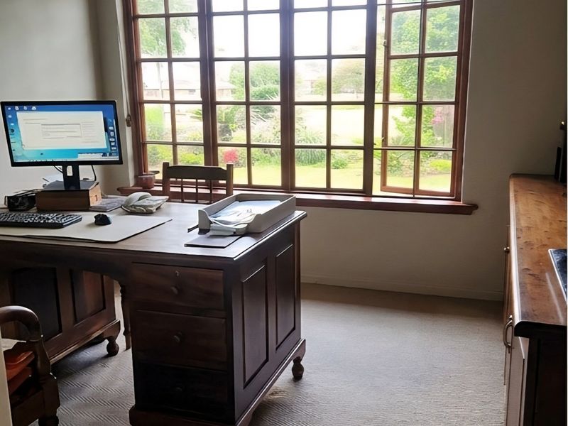 A well-lit home office featuring timber-framed windows with garden views, neutral carpeting, and built-in cabinetry.
