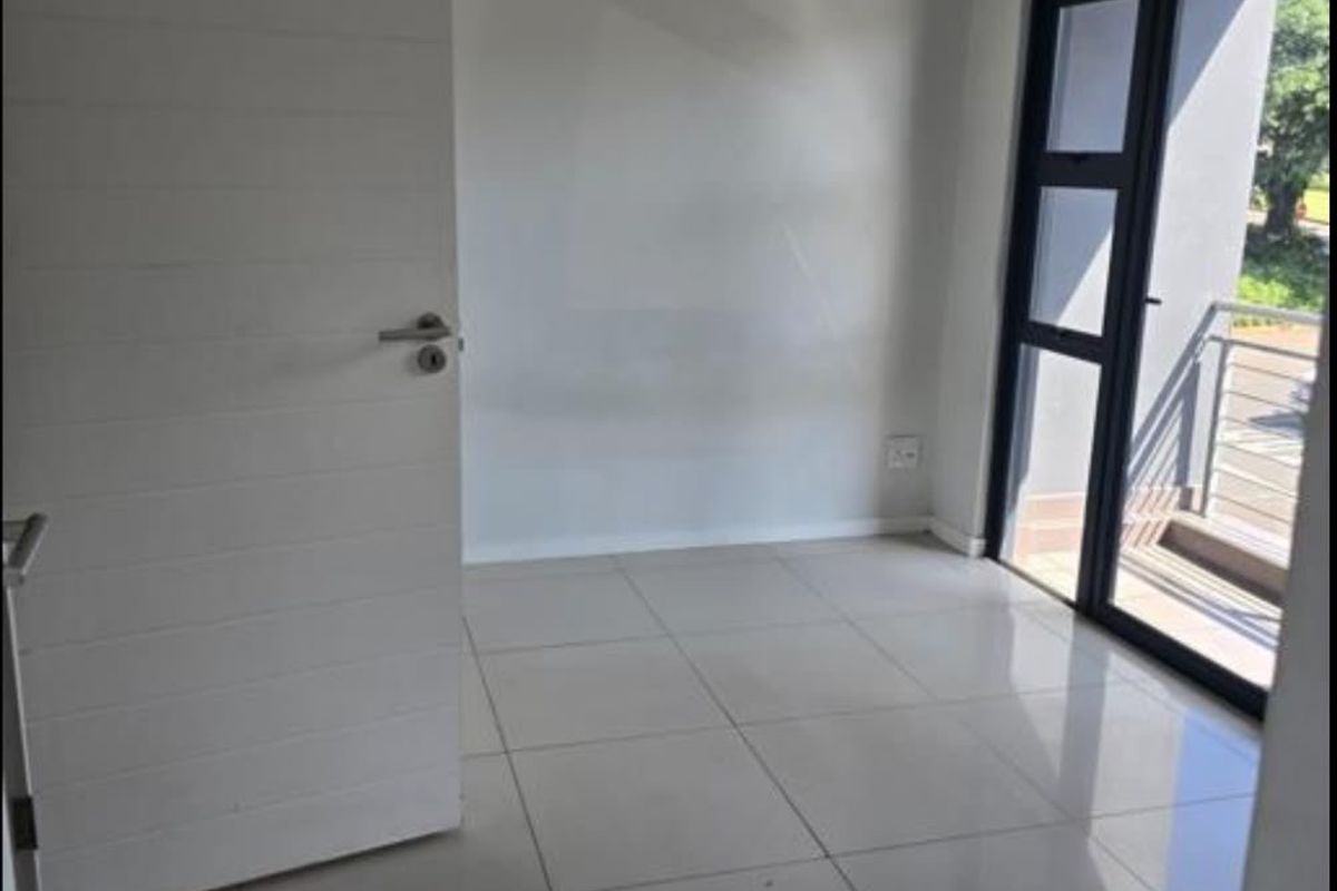 A bright, empty bedroom featuring neutral tiled flooring and a modern aluminium-framed glass door providing direct access to a private balcony.