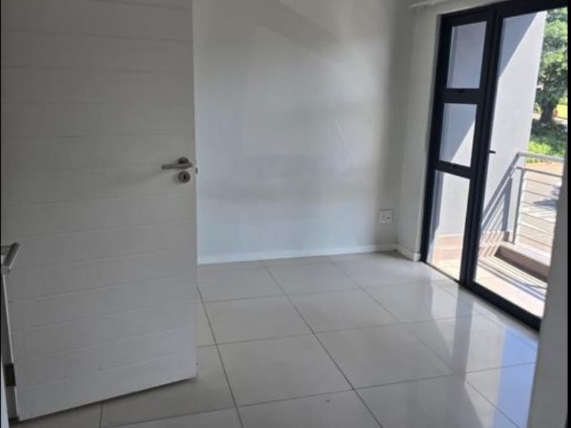 A bright, empty bedroom featuring neutral tiled flooring and a modern aluminium-framed glass door providing direct access to a private balcony.