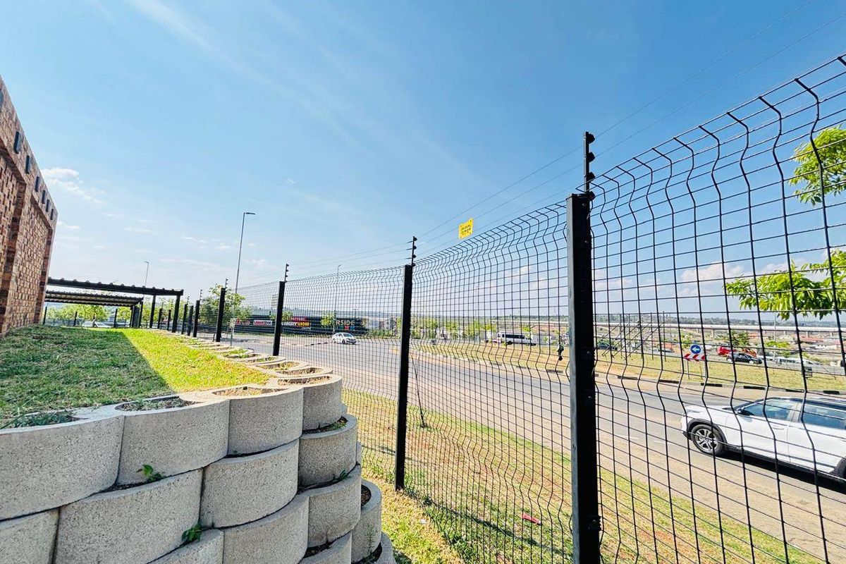 Exterior view of the property boundary featuring a retaining wall, mesh security fencing with electric wires, and a brick building facade.