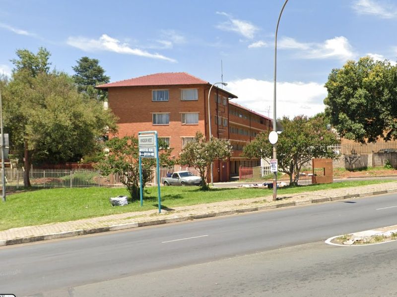 Street view of a multi-story brick apartment building with a tiled roof, featuring perimeter fencing and access from the main road.
