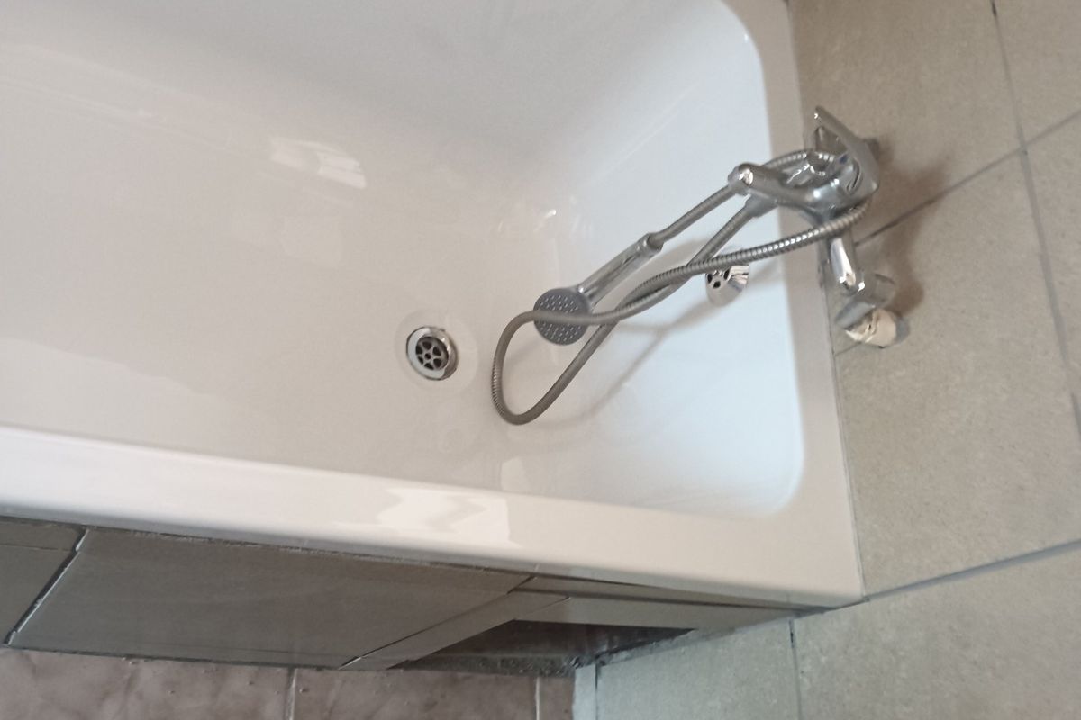 A compact bathroom featuring a bathtub with a handheld shower mixer, tiled walls, and flooring, requiring some maintenance to the tile finishes.