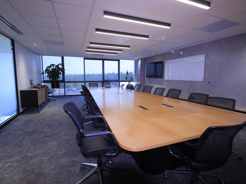 Modern professional boardroom featuring a large central table, floor-to-ceiling windows with natural light, integrated lighting, and a sleek concrete-effect feature wall.