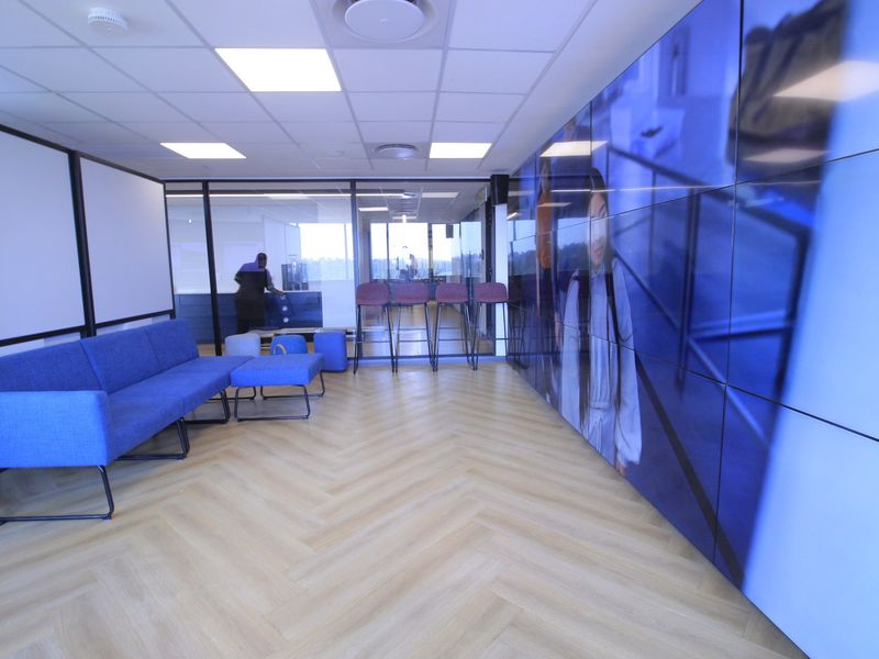 Modern commercial office space featuring herringbone flooring, glass partitions, and a large integrated digital display wall, creating a contemporary professional environment.