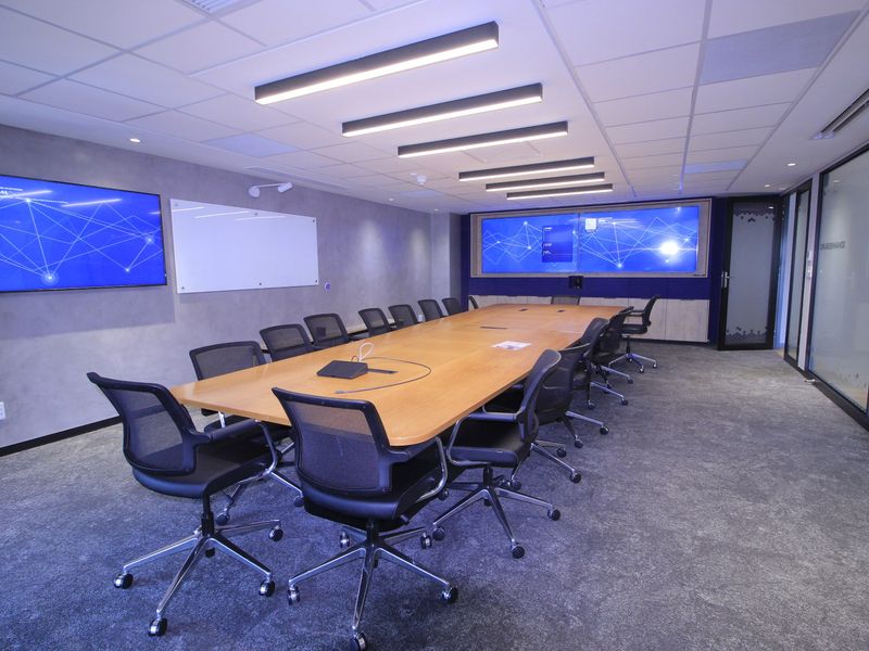 Modern professional boardroom featuring a large conference table, integrated wall-mounted display screens, glass partitions, and recessed linear lighting in a suspended ceiling.