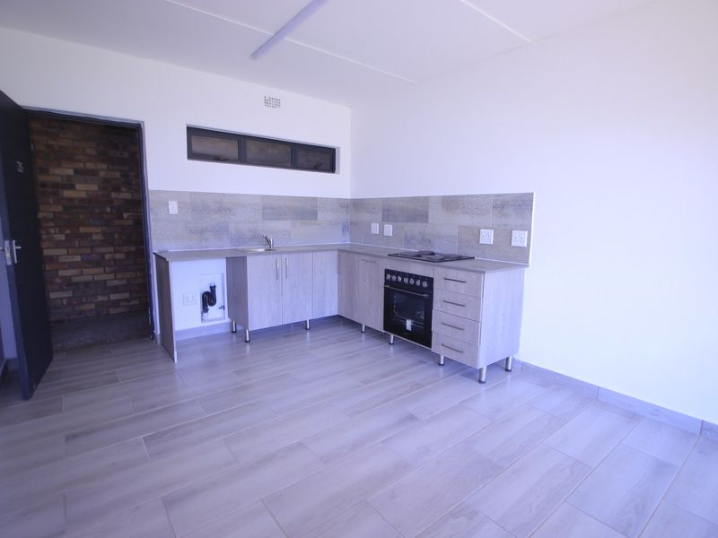 A clean, modern kitchen featuring light wood-finish cabinetry, built-in oven and hob, tiled backsplash, and neutral floor tiles in an open-plan layout.
