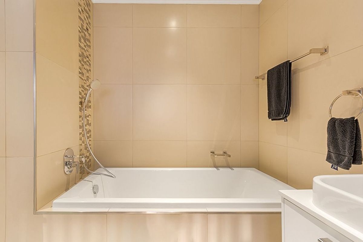 A clean bathroom featuring a built-in bathtub with a handheld shower head, complemented by neutral wall tiles and a decorative mosaic accent strip.
