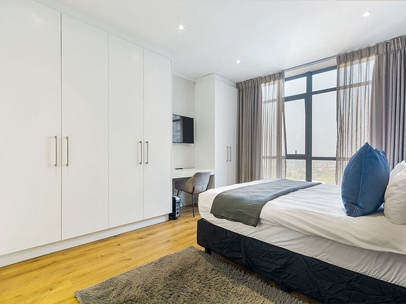A modern, well-lit bedroom featuring extensive built-in cabinetry, a dedicated desk area, hardwood flooring, and large windows offering expansive views.