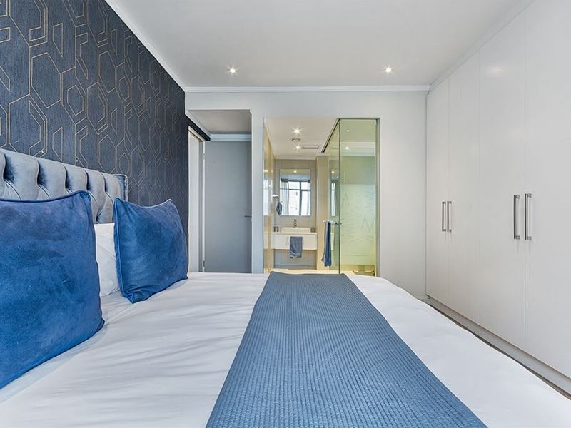 Modern bedroom featuring built-in cupboards, recessed lighting, and an integrated en-suite bathroom with a glass shower enclosure and vanity basin.
