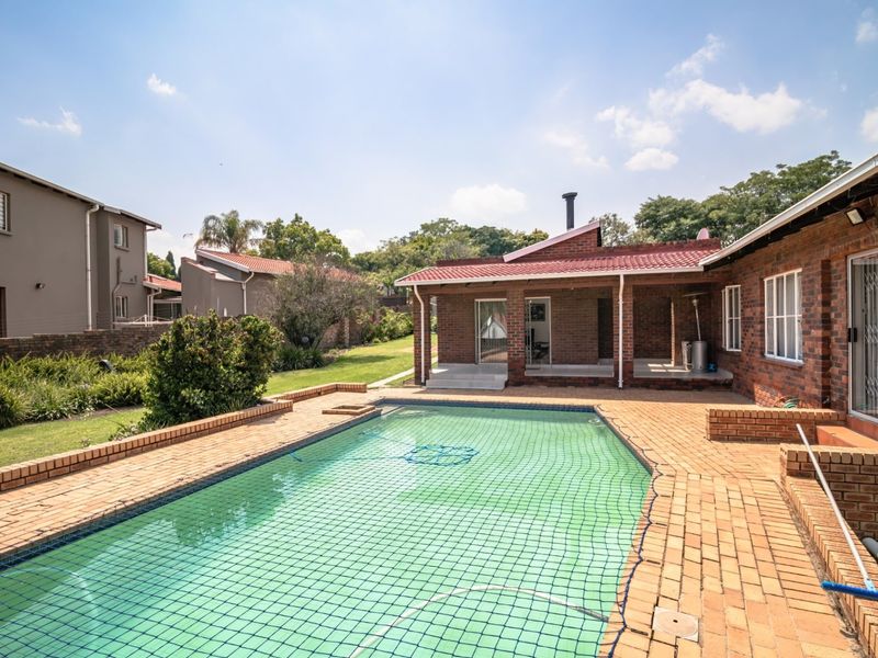 A spacious backyard featuring a swimming pool with safety net, brick-paved surround, and a covered patio area attached to the main brick residence.