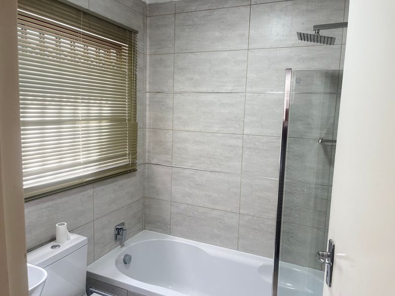 A clean, modern bathroom featuring a built-in bathtub, glass shower screen, overhead shower, and neutral wall tiling.