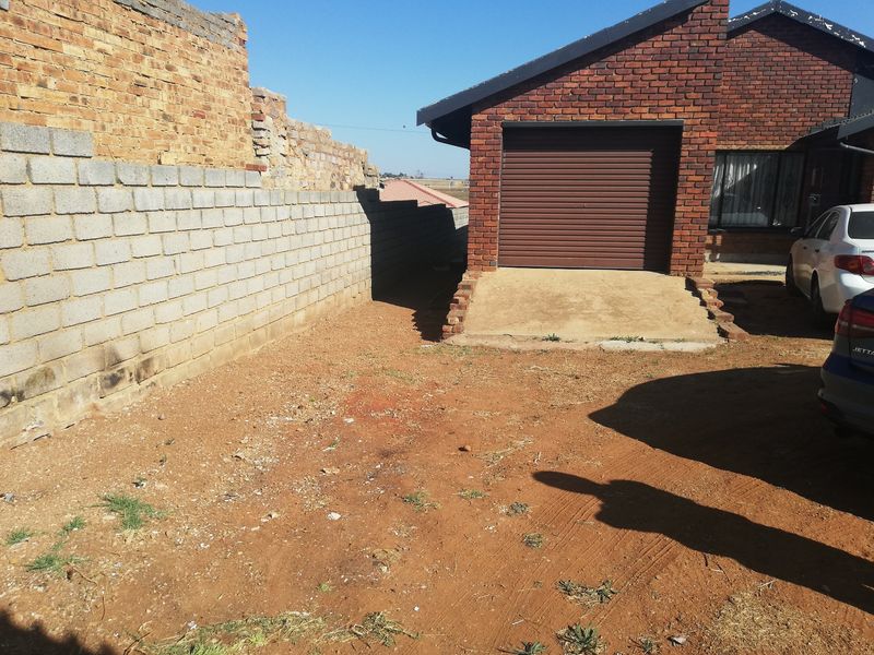 Front exterior view showing a face brick house with a roller shutter garage door and an unfinished boundary wall on a dirt driveway.