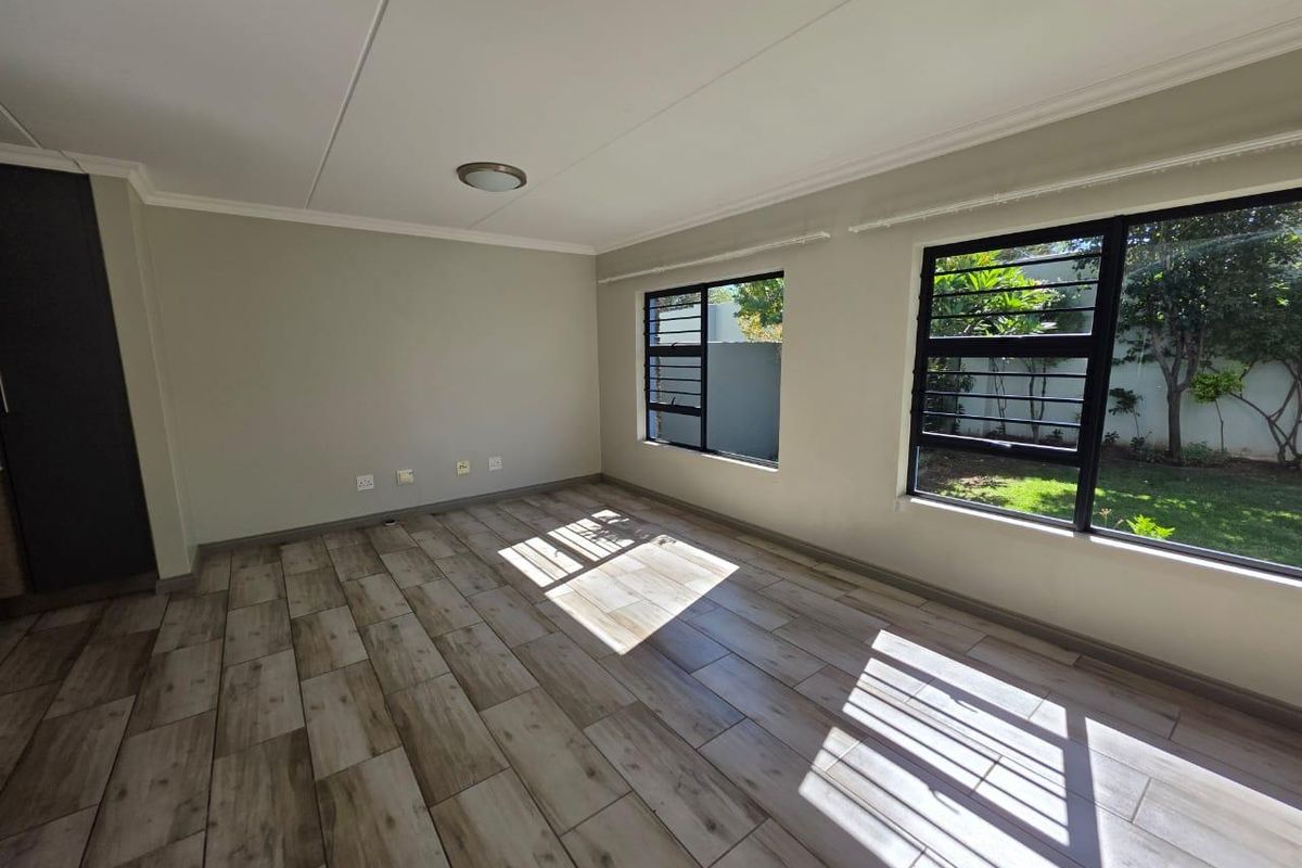 A bright, empty living room featuring modern wood-look tiled flooring, large windows with security bars, and a neutral color palette.