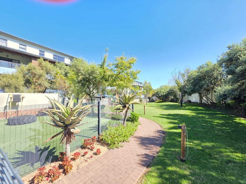 A well-maintained communal garden area featuring a paved walkway, manicured lawn, and secure perimeter fencing within the apartment complex grounds.