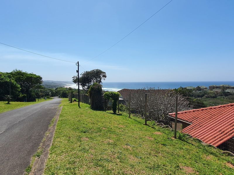 A street-level view of the residential property showing the access road, perimeter fencing, and a scenic coastal landscape in the background.