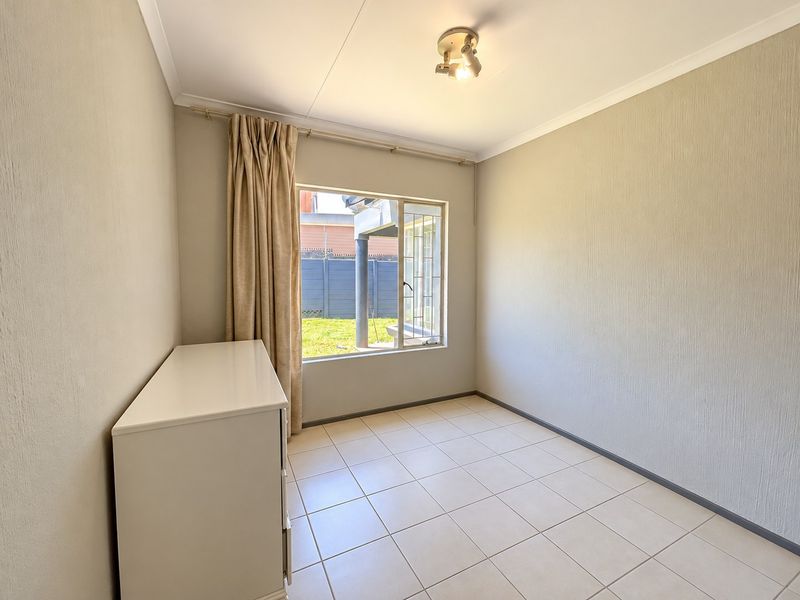 A bright, empty bedroom featuring neutral tiled flooring, clean wall finishes, and a window with security bars overlooking the rear garden area.