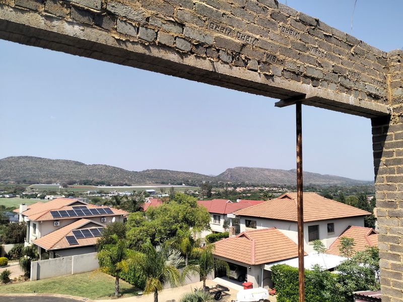 An elevated view from an unfinished construction site showing a concrete lintel and metal support column overlooking a residential neighborhood with mountain views.