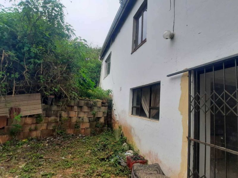 Rear exterior view of the property showing a side wall, security gate, and a retaining wall with significant overgrown vegetation and weathered paintwork.